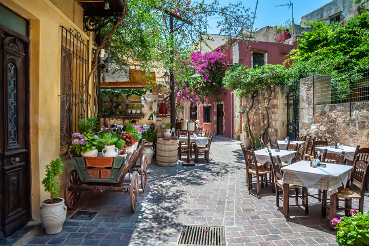 Charming Greek alley with outdoor café tables, colorful flowers, and stone-paved streets creating a warm Mediterranean atmosphere.