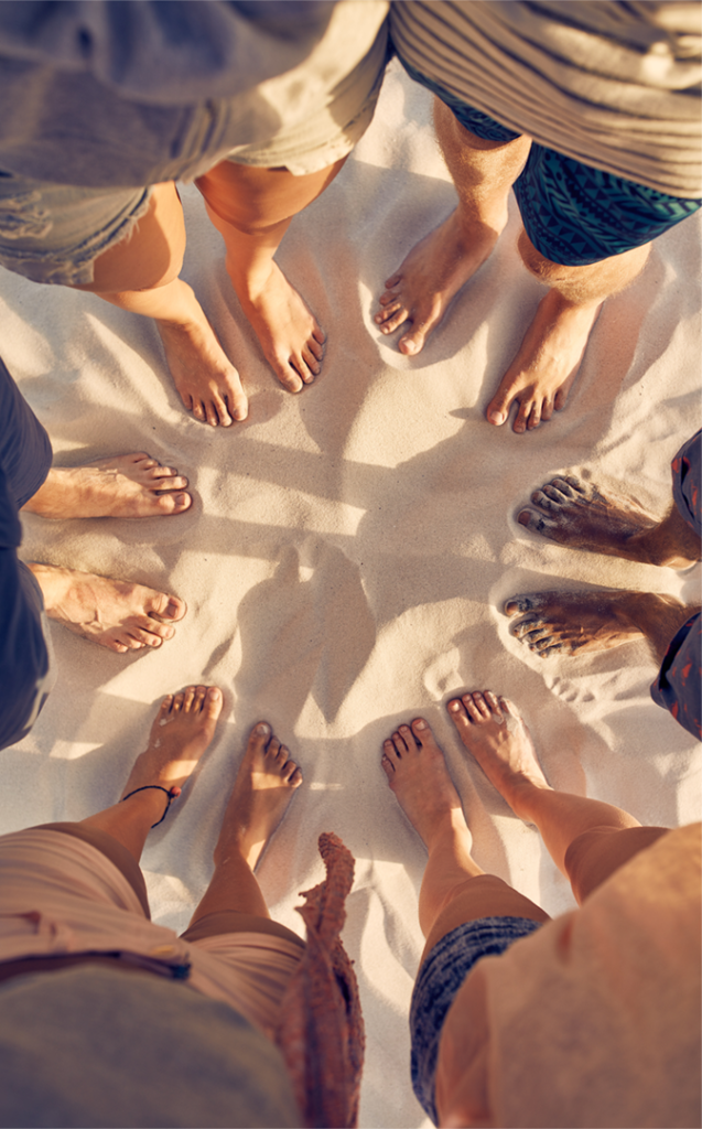 Group of friends standing barefoot in a circle on a sandy beach, showing togetherness and summer vibes.