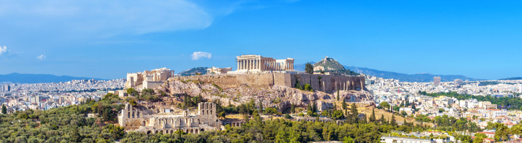 Panoramic view of the Acropolis in Athens with the Parthenon and surrounding cityscape, overlooking the Aegean Sea and distant mountains under a clear blue sky.