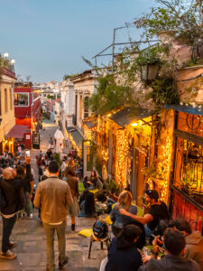 Evening view of a lively narrow street in Plaka, Athens, filled with people dining outdoors, surrounded by colorful buildings and warm string lights.