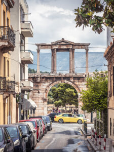 Street view of Hadrian’s Arch in Athens, Greece, framed by residential buildings and parked cars, with mountains visible in the background.