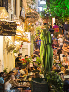 Evening view of a lively narrow street in Plaka, Athens, filled with people dining outdoors, surrounded by colorful buildings and warm string lights.