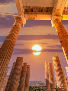 Dramatic sunset view framed by ancient marble columns of a Greek temple, with the glowing sun above the city of Athens and warm golden light illuminating the ruins.
