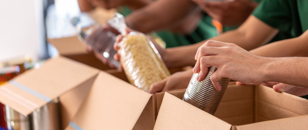 emergency-relief Close-up of people packing food items like canned goods, pasta, and bottled water into cardboard boxes for donation.