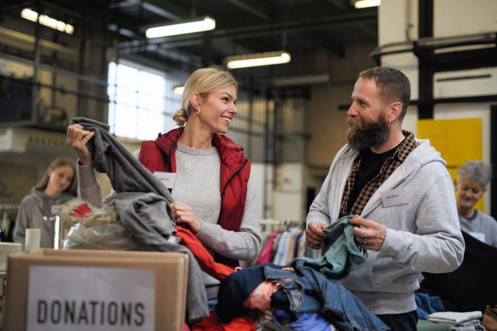 Volunteers smiling as they sort and fold clothes at a donation center, surrounded by boxes labeled “Donations.”