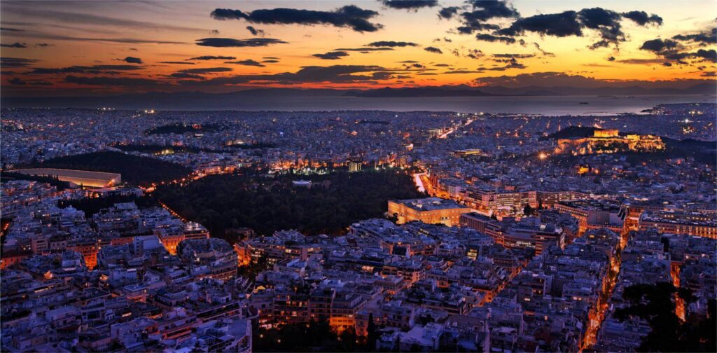 A panoramic view of Athens at sunset, with the Acropolis illuminated and city lights glowing under a colorful sky.