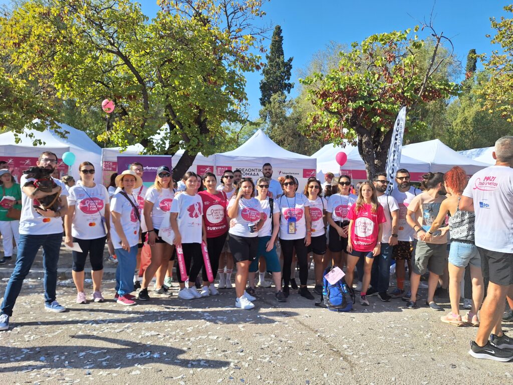 Group of TP volunteers participating in the Greece Race for the Cure event, wearing pink and white shirts with breast cancer awareness messages. They are smiling, holding banners and balloons, celebrating solidarity and support for the cause.