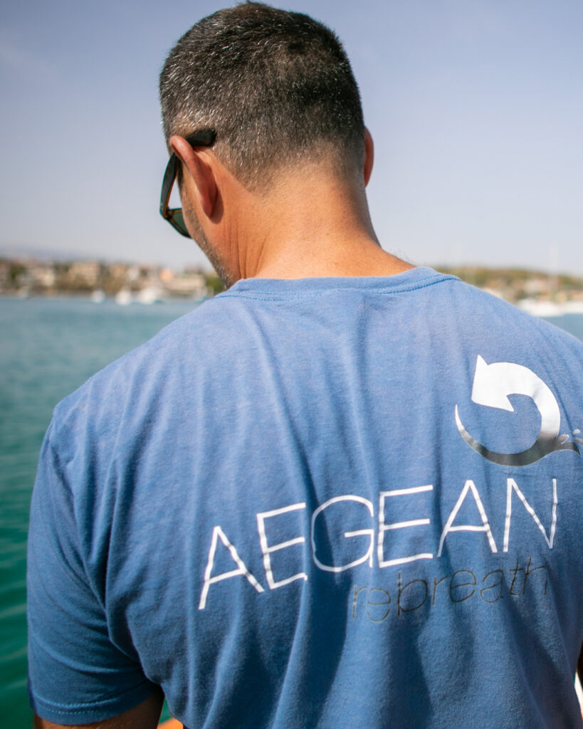 Person wearing a blue “AEGEAN rebreath” T-shirt, standing near the sea with boats and a coastal town visible in the background.