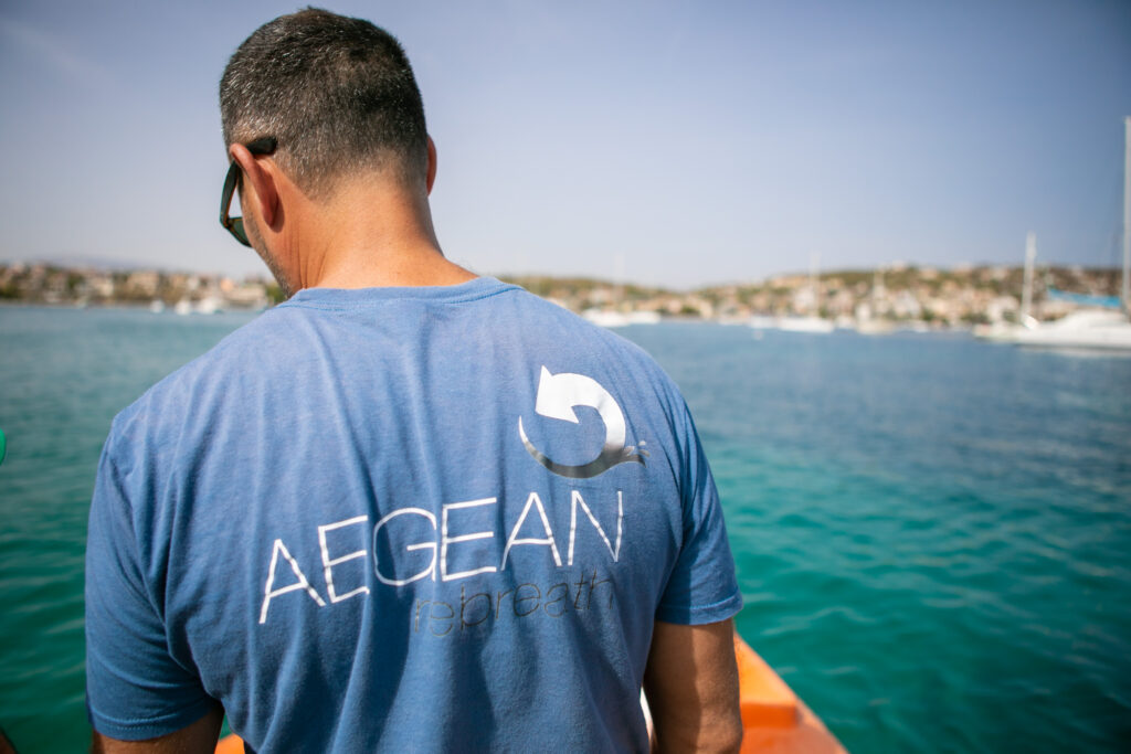 Person wearing a blue “AEGEAN rebreath” T-shirt, standing near the sea with boats and a coastal town visible in the background.