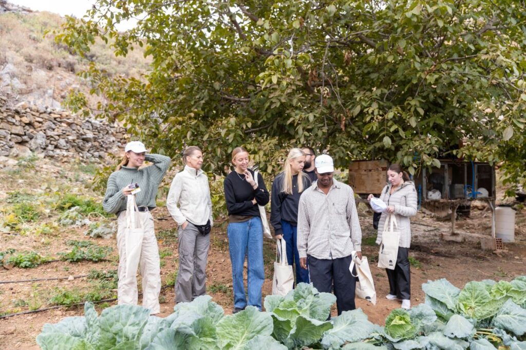 Group of people standing outdoors in a garden or farm area with green leafy plants in the foreground, some holding tote bags and notebooks, while listening and smiling under a large tree.