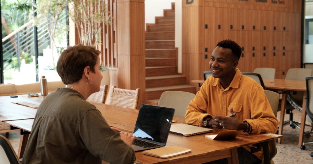 Two people sitting at a wooden table in a modern office having a friendly conversation, with laptops open in front of them.
