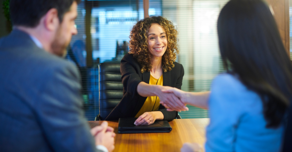 Smiling professional woman in a business meeting shaking hands with another person across the table, while a colleague looks on.
