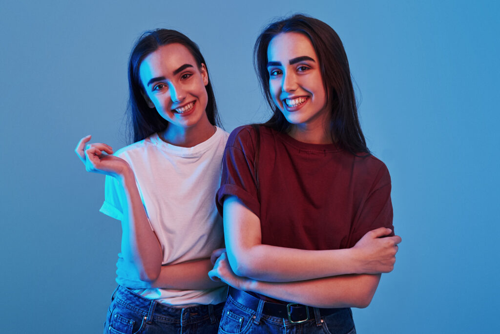 Two smiling women who look alike standing close together against a blue background, one wearing a white shirt and the other a red shirt, posing confidently.