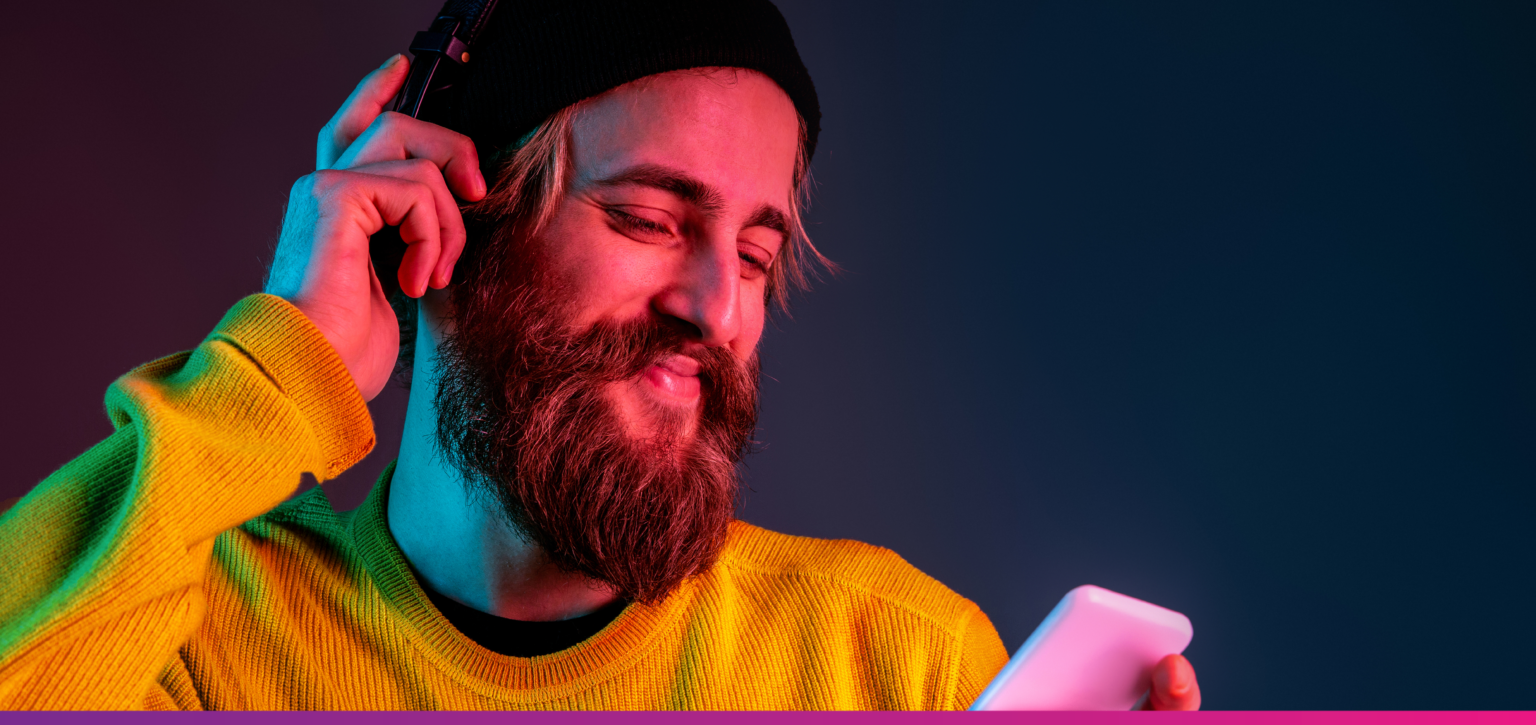 A man wearing a yellow sweater and headphones, smiling while listening to music and looking at his phone under colorful neon lighting.