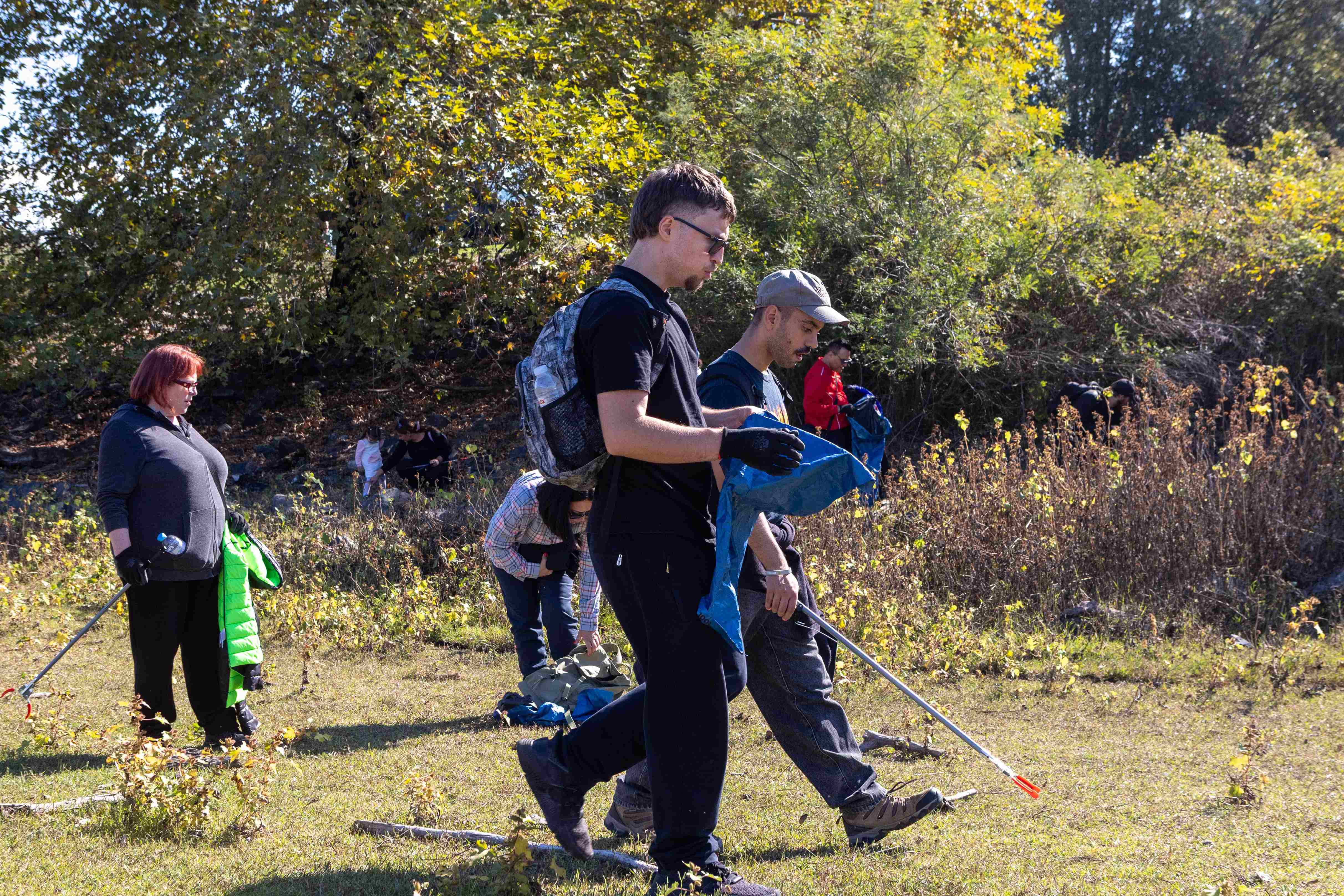 TP Volunteers cleaning up at Thessaloniki at lake kerkini, picking up litter with grabbers and bags, surrounded by trees on a sunny day.