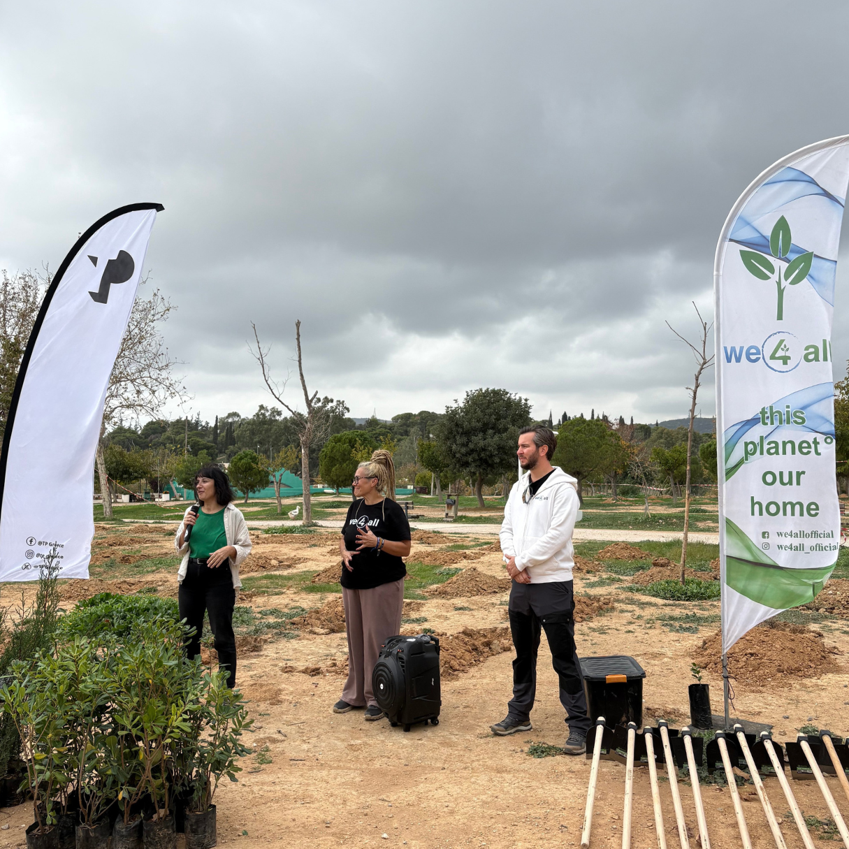 We4all Speakers addressing volunteers during a tree-planting event, standing between two event banners with potted plants and tools arranged on the ground.