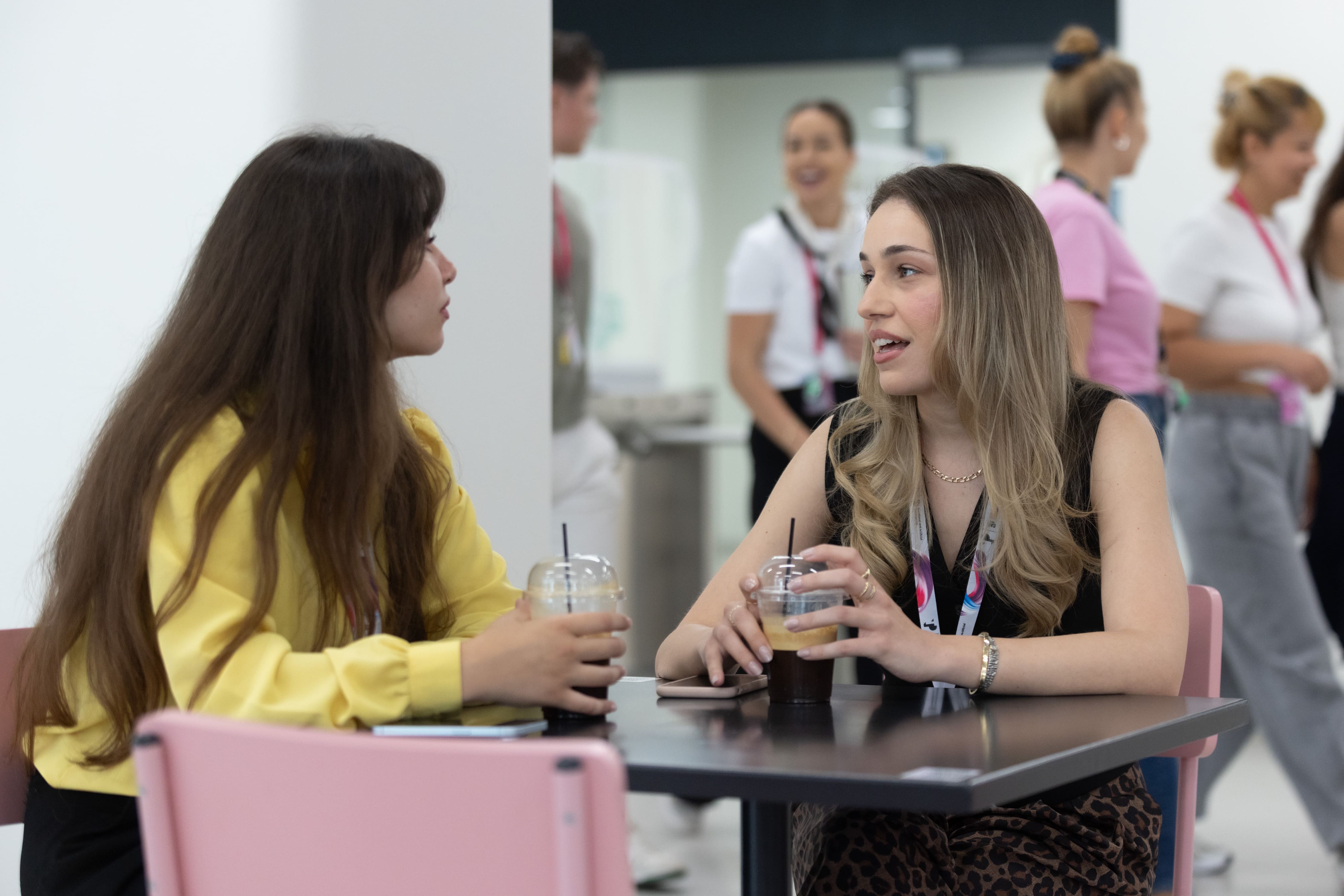 Two women sit at a table in a bright office break area, talking while holding iced coffees. Several colleagues can be seen chatting and walking in the background.