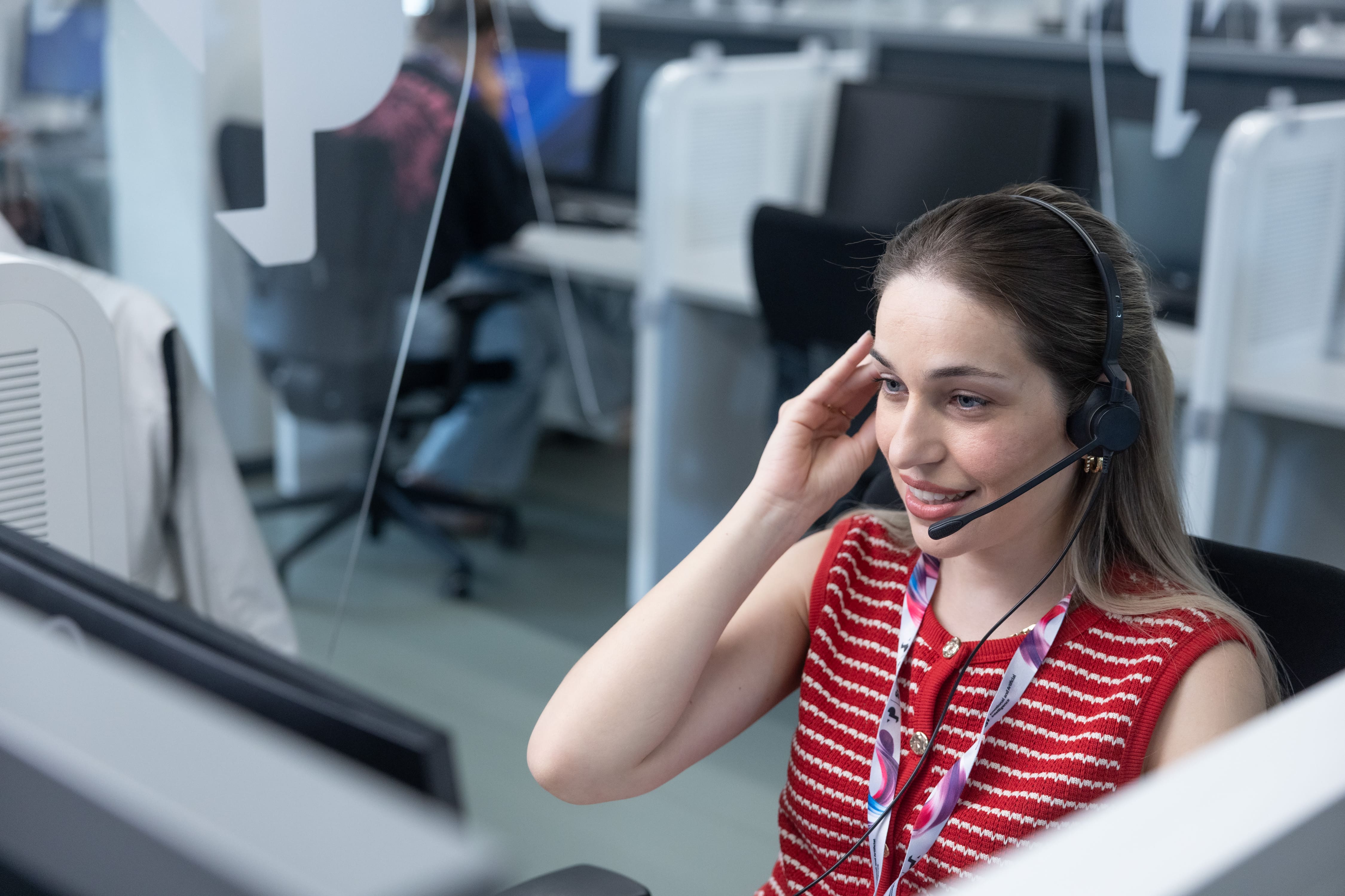 A woman wearing a headset sits at a workstation in a modern office, smiling as she takes a call. She is surrounded by computer monitors, with other employees working in the background.