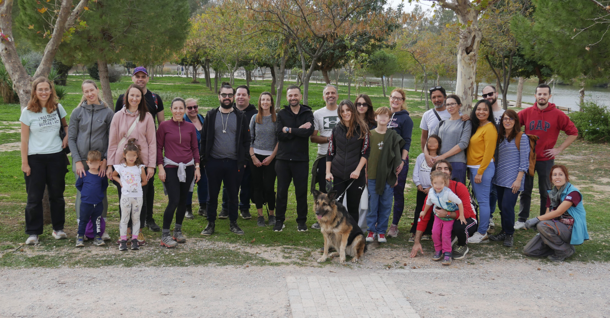Large group of adults and children posing together outdoors in a park, standing in front of trees and greenery. A dog sits at the front of the group. Everyone appears relaxed and smiling, suggesting a friendly gathering or community event.