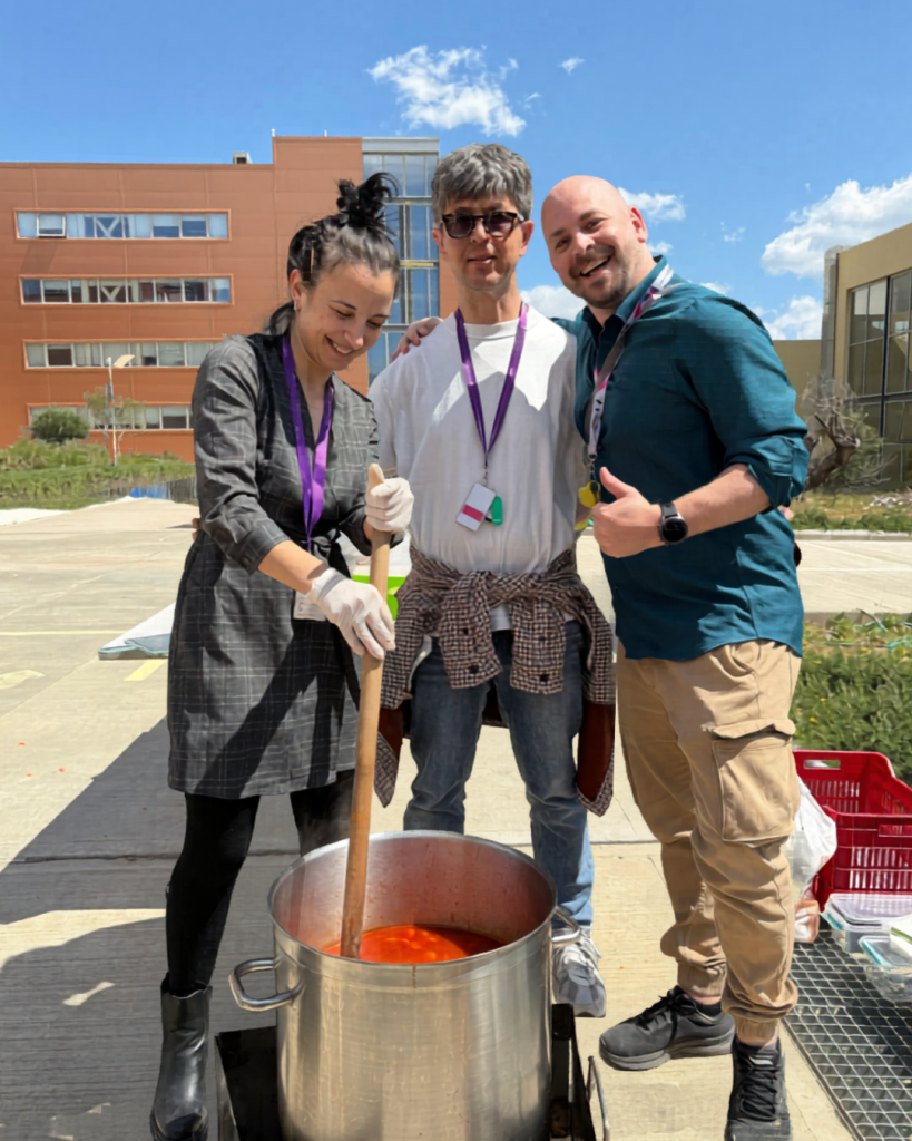 Three TP Greece employees smiling while preparing food outdoors. One person stirs a large pot of red soup or sauce, while the others stand beside them, wearing lanyards and casual clothing. The scene is sunny, with modern office buildings in the background, conveying teamwork and community impact.