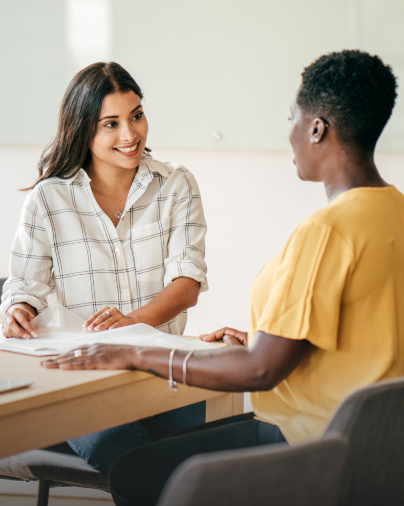 Two people seated at a desk having a friendly, professional conversation, with one person showing documents and smiling while the other listens attentively.