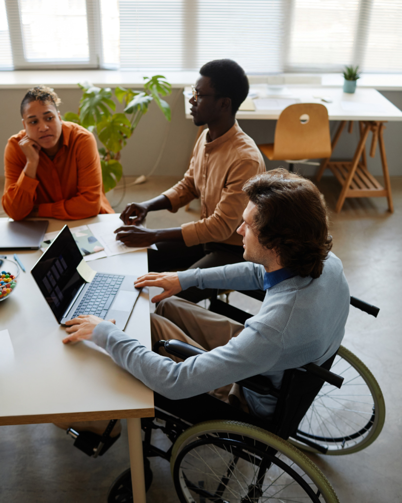 Three colleagues collaborate around a desk in a bright office, including a man using a wheelchair working on a laptop while two coworkers discuss ideas nearby.