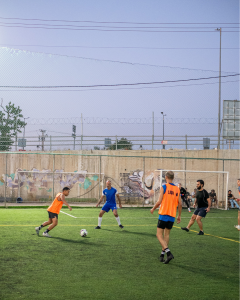 Several adults play a small-sided soccer game on an outdoor artificial turf field. One player in an orange training bib dribbles the ball while teammates and opponents in blue, black, and orange gear position themselves around him. A goal stands in the background, with chain-link fencing, graffiti-covered walls, utility poles, and a clear evening sky visible beyond the field.