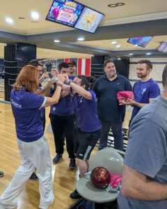 TP Greece team members celebrate a bowling competition, raising a trophy together at a bowling alley while colleagues applaud.