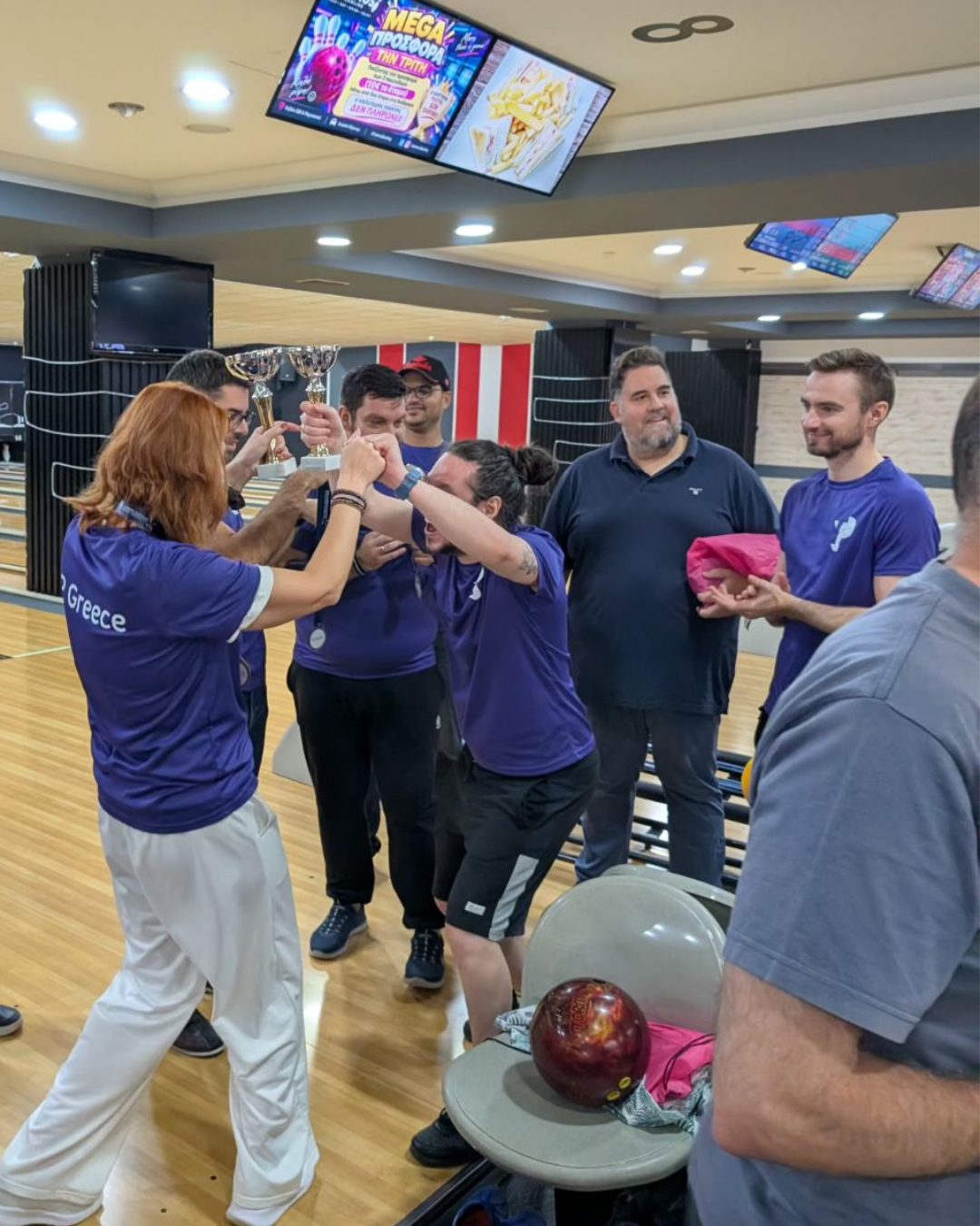 TP Greece team members celebrate a bowling competition, raising a trophy together at a bowling alley while colleagues applaud.